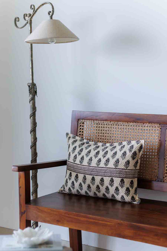 A block print lumbar cushion with a black and green pattern rests on a wooden bench with a woven cane backrest, next to a metal floor lamp.