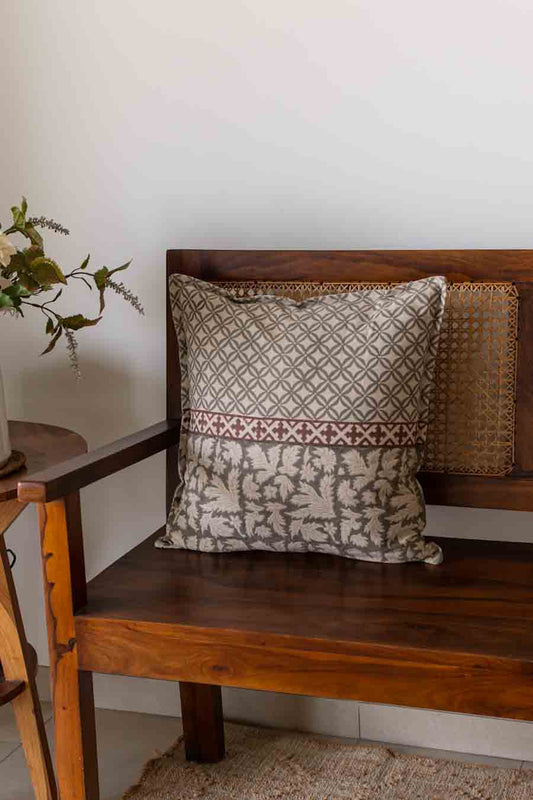 A square decorative cushion with a grey and rust geometric and floral pattern, resting on a wooden bench.