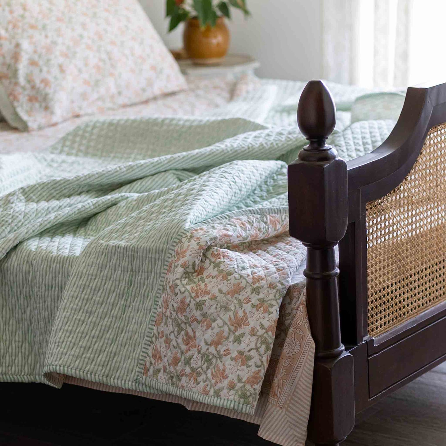 A quilted bedding set styled on a bed with a dark wood cane headboard. The set features a sage green and white striped quilt, a matching striped pillowcase, and a floral print quilt in soft pink, peach, and green tones.