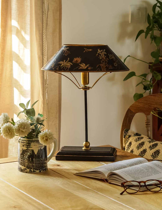 A table lamp with a black and gold floral shade and brass-toned metal base, styled on a wooden table next to an open book and glasses