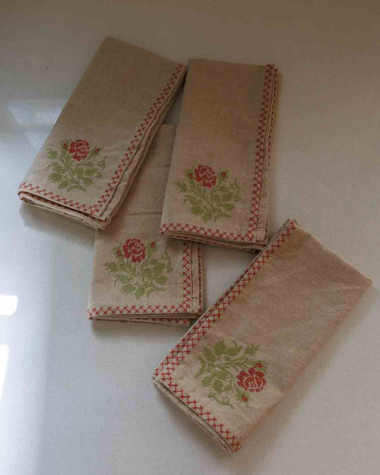 Four beige cotton embroidered napkins with red stitched borders and green floral motifs, arranged on a light-colored surface.