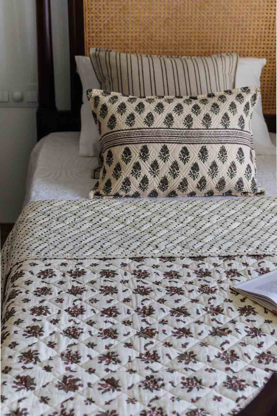A styled bed featuring a cream-colored quilt with a brown floral pattern, a matching patterned pillow, and striped pillows against a woven cane headboard.