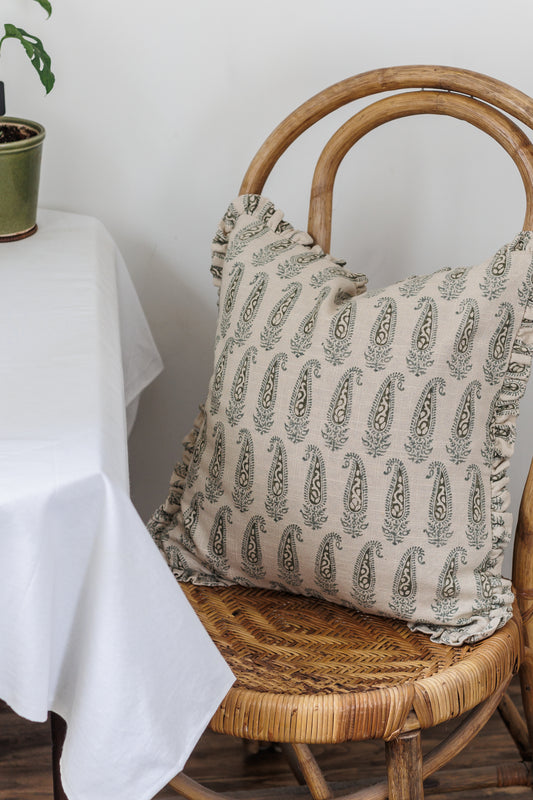 A cushion cover with a paisley block print pattern in shades of green, featuring ruffles and placed on a wicker chair with a table and plant