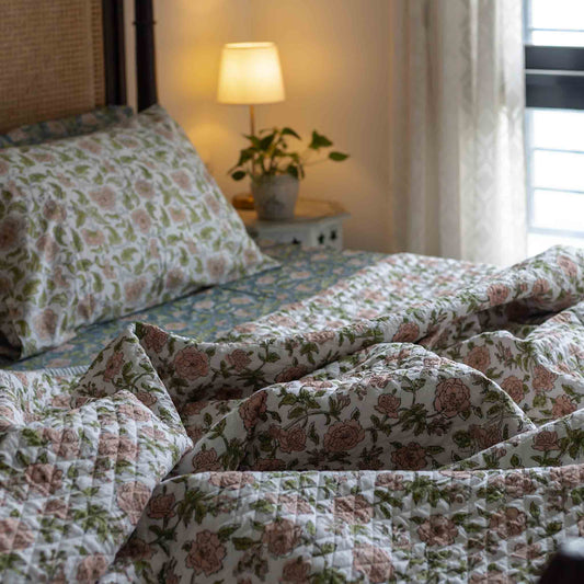 A quilted bedding set with a vintage floral pattern in dusty pink and sage green on a cream background, styled on a bed in a warmly lit bedroom.