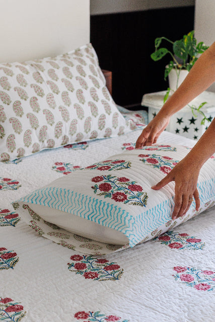 A quilted cotton bedspread with hand block print featuring floral patterns, placed on a bed with two pillows, in a bedroom with a a potted plant in the background.
