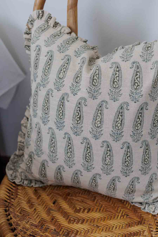 A cushion cover with a paisley block print pattern in shades of green, featuring ruffles and placed on a wicker chair.