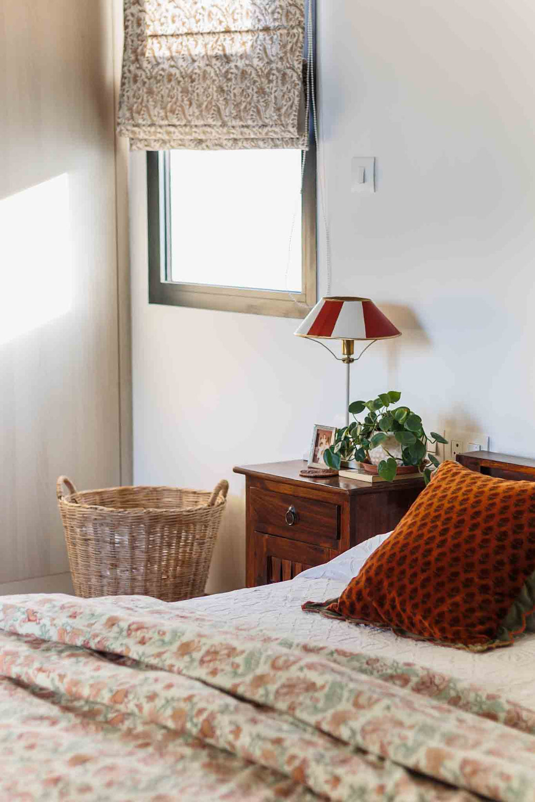 A bedroom with quilt , red pillows, table lamp , wicker basket next to a window