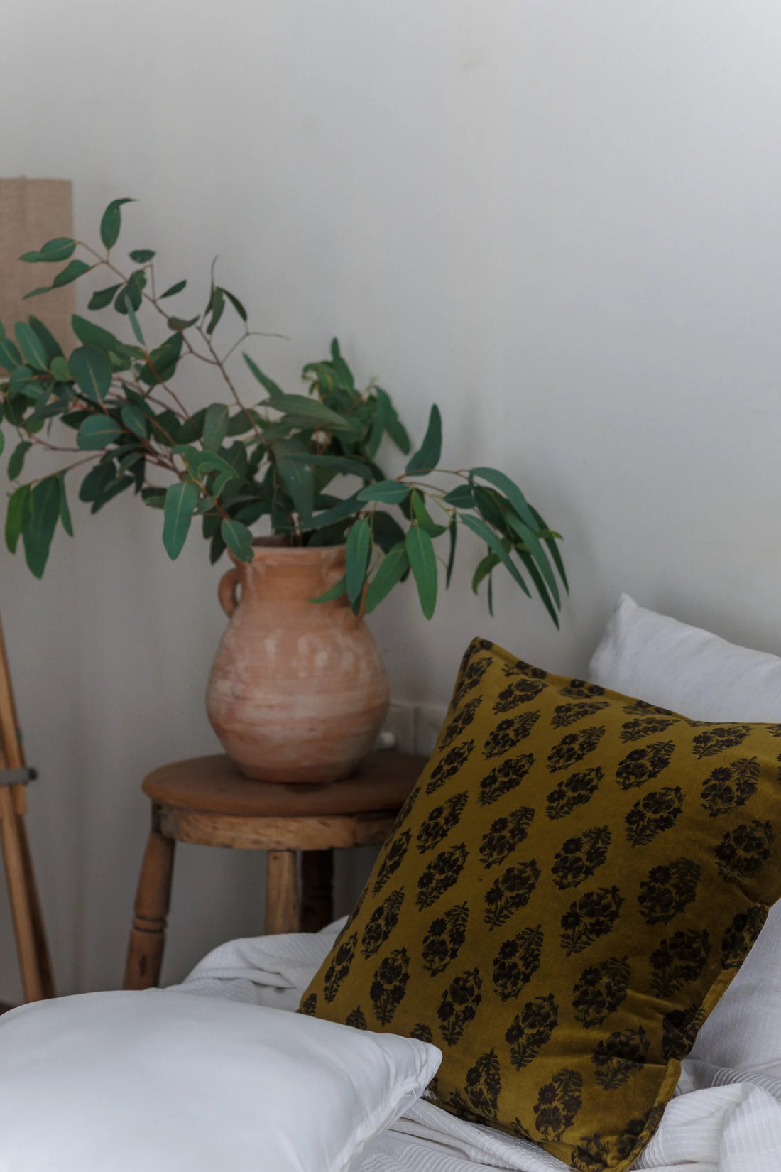 A green block print cushion on white bedding with Vase and green leaves