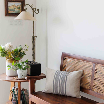 Wooden bench with wicker backrest and striped linen pillow, next to a small table with plants and lamp.