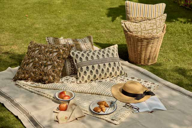 A styled outdoor picnic setup on a beige blanket , featuring patterned throw pillows, a woven basket of cushions, a straw hat , a bowl of fruit, and an open book.