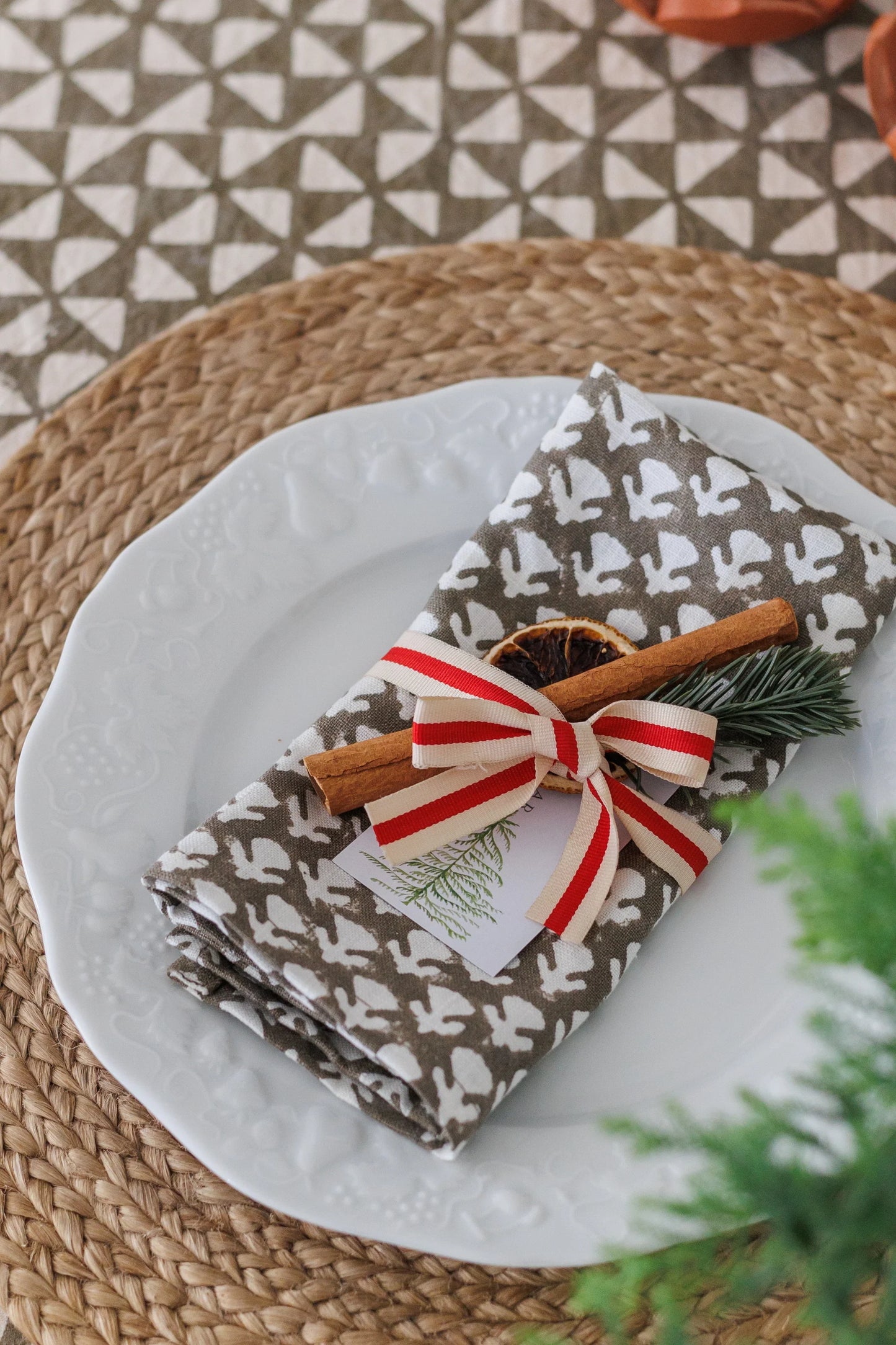 Folded napkin with block print pattern on a plate with cinnamon stick and pine sprig, on a textured surface.
