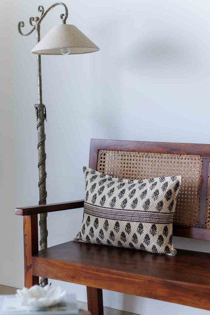 A block print lumbar cushion with a black and green pattern rests on a wooden bench with a woven cane backrest, next to a metal floor lamp.