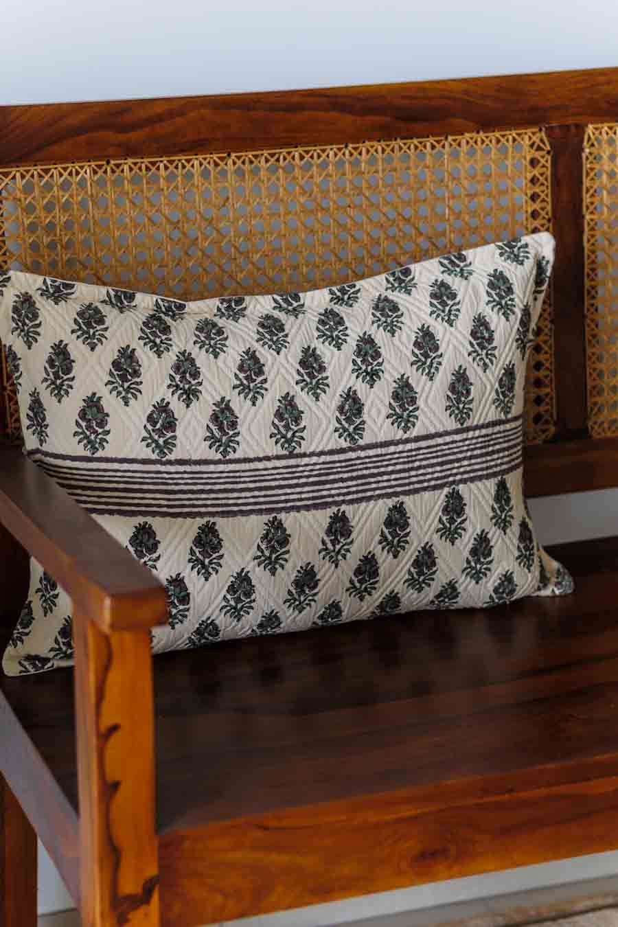 A rectangular throw pillow with a black and off-white block print pattern, resting on a wooden bench with a woven cane backrest.