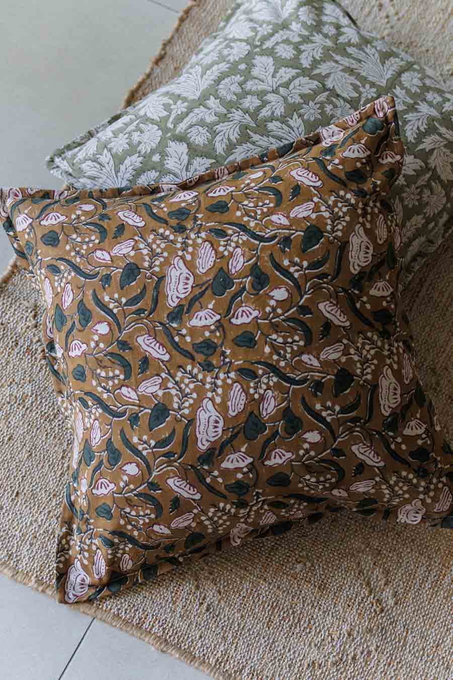 A square decorative throw pillow with a brown, beige, and green botanical block-print pattern, styled on a woven jute rug with another patterned pillow in the background.