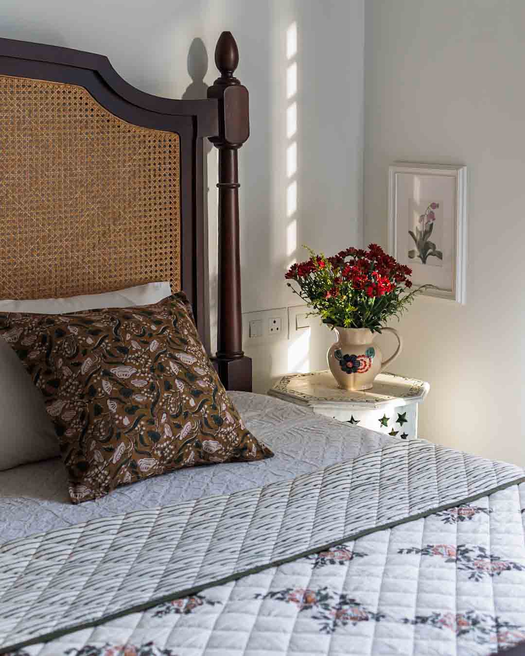 A styled bedroom with cane headboard, a Cinnamon block print cushion cover with a quilted floral quilt. 