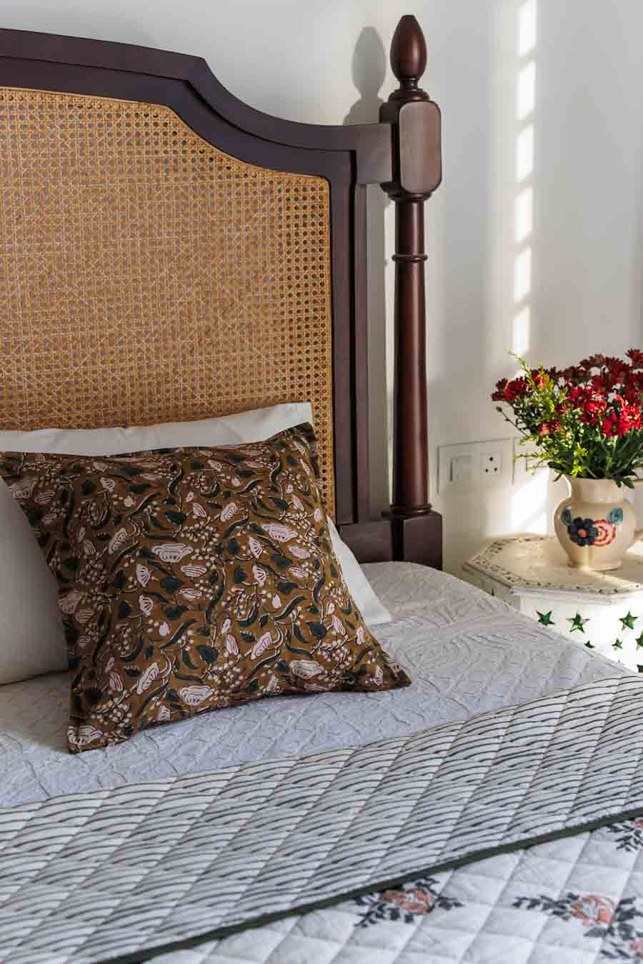 A styled bedroom scene featuring wood and woven cane headboard, a patterned brown accent pillow, and a quilted green bedspread.