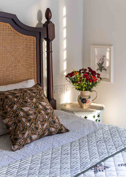 A bedroom featuring a cane headboard, a block print cushion cover with a quilted bedspread, a small bedside table holds a red flowers in a vase.