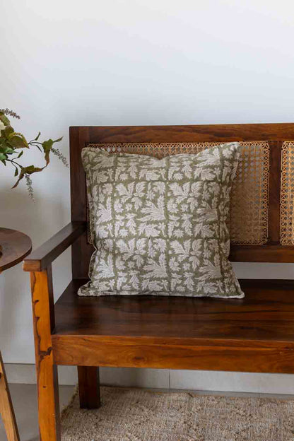 A square cushion with a beige and olive green botanical pattern, resting on a wooden bench with woven cane backrest.