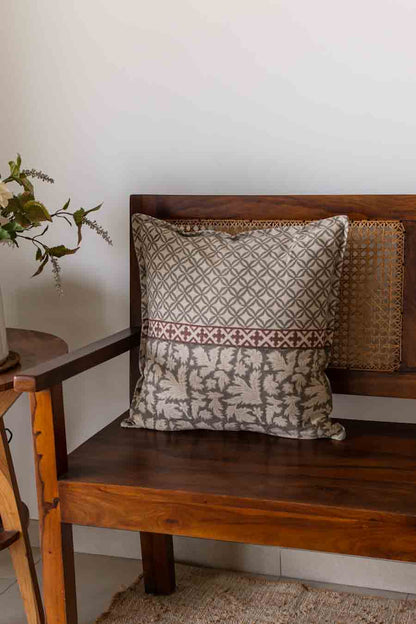 A square decorative cushion with a grey and rust geometric and floral pattern, resting on a wooden bench.