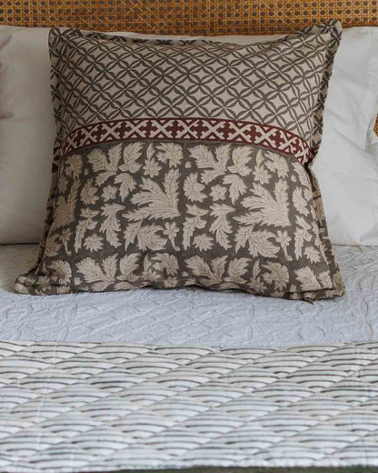 A printed cushion cover in grey botanical damask pattern and a red geometric border, styled on a bed with a textured quilt.
