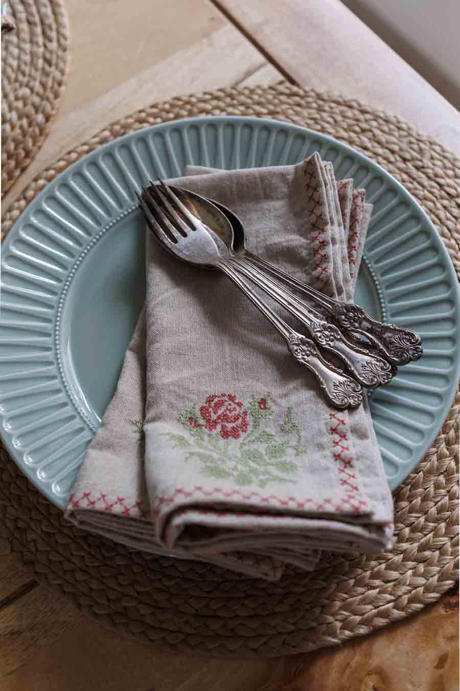 A folded linen napkin with a pink rose and green floral embroidery sits on a light blue plate, topped with tsilver cutlery