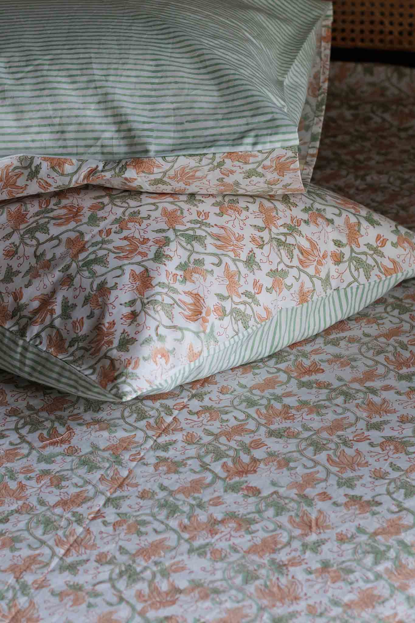 A close-up of patterned bedding with matching pillows, featuring a white base with green stripes on the top pillow and a floral print in peach and green across the quilt and other pillows.