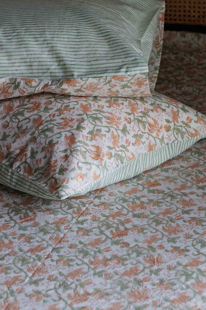 A close-up of patterned bedding with matching pillows, featuring a white base with green stripes on the top pillow and a floral print in peach and green across the quilt and other pillows.