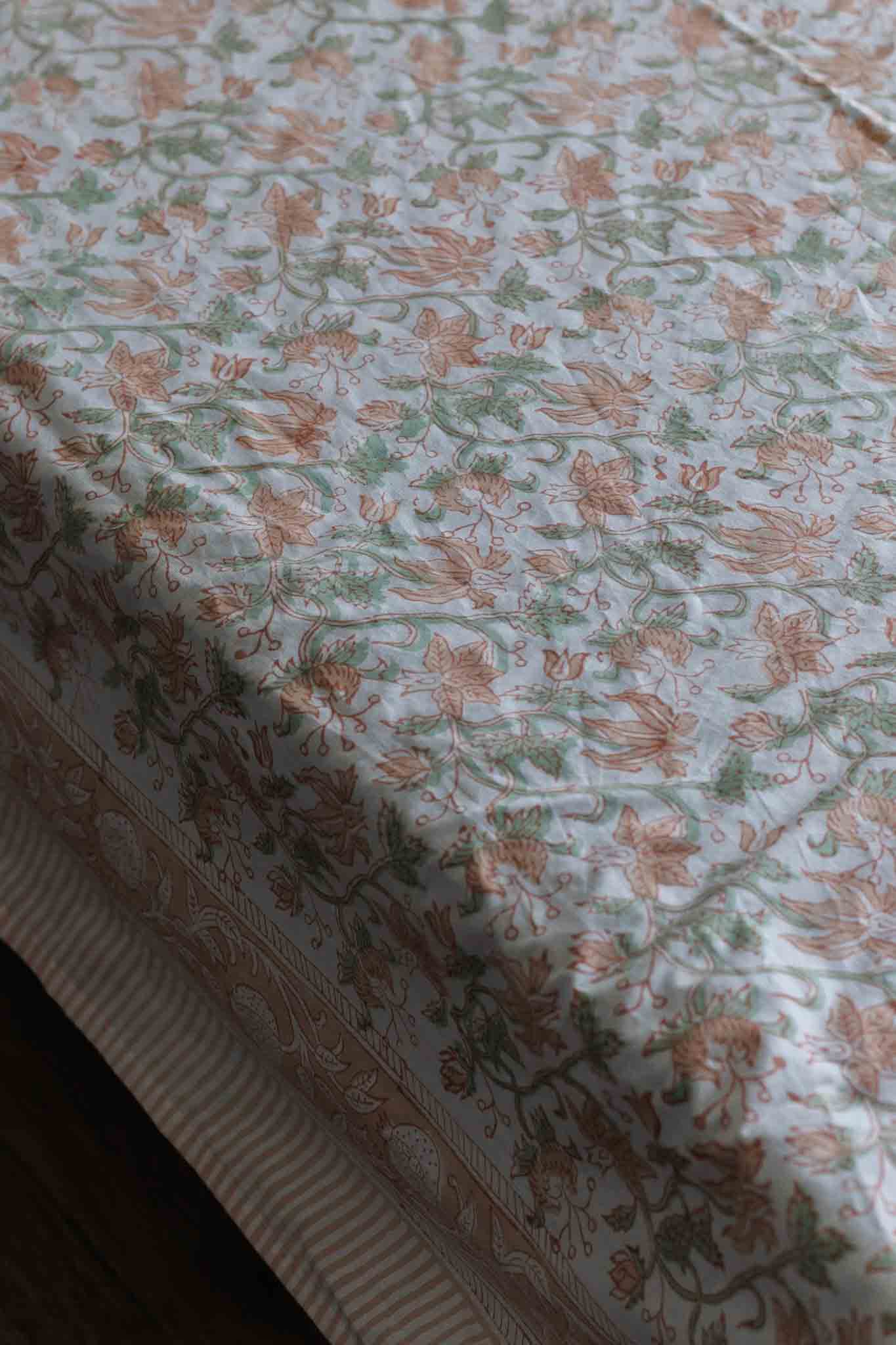 A close-up of a block print bed sheet with a delicate floral and botanical pattern in muted peach, sage green, and cream on a white background.