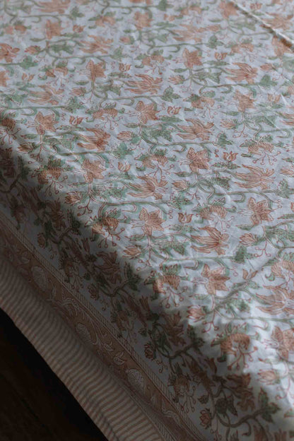 A close-up of a block print bed sheet with a delicate floral and botanical pattern in muted peach, sage green, and cream on a white background.