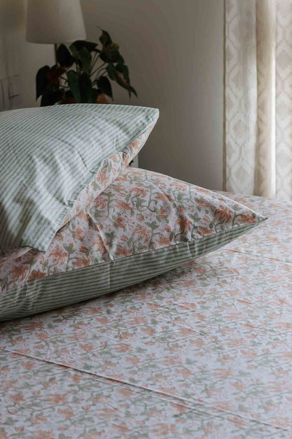 A close-up of a bed styled with matching bedding, featuring a green and white striped pillowcase with a white floral insert, a striped pillow, and a floral sheet in soft peach, green, and white tones.