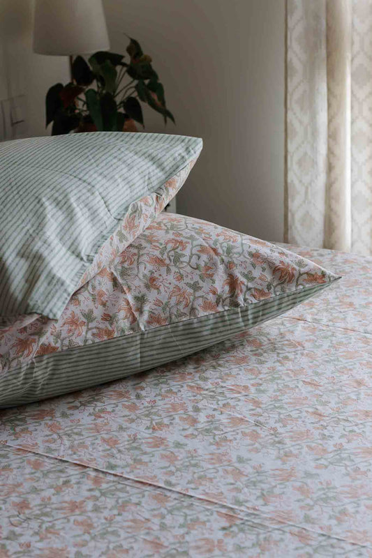 A close-up of a bed styled with matching bedding, featuring a green and white striped pillowcase with a white floral insert, a striped pillow, and a floral sheet in soft peach, green, and white tones.