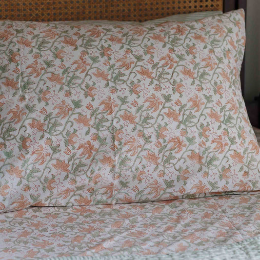 A close-up of a floral-patterned pillowcase and matching bed sheet with peach and sage green flowers on an off-white background, styled in a bedroom setting with a woven cane headboard.