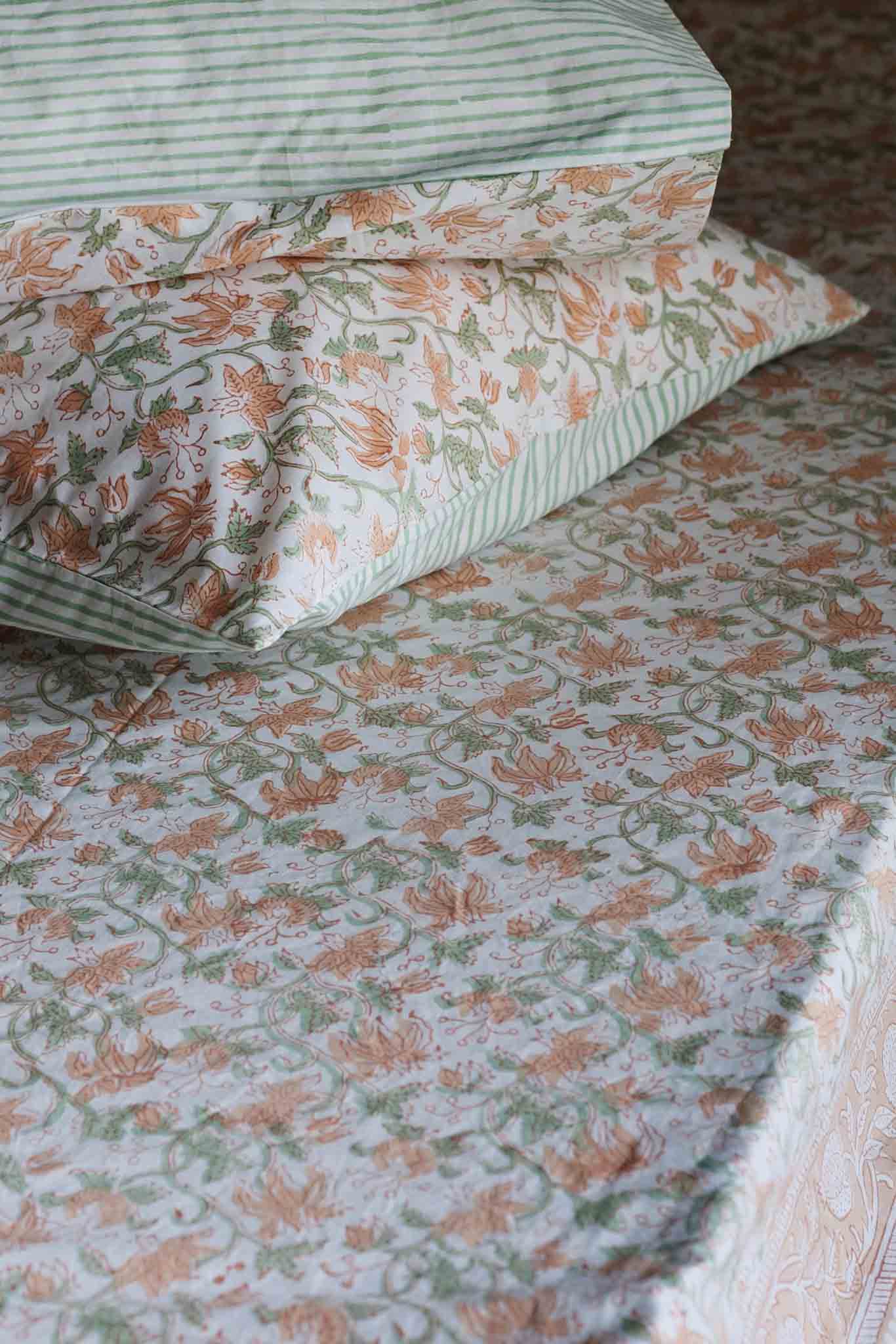 A close-up of a bed dressed in a floral print sheet and matching pillowcases. The pattern features orange flowers and green leaves on a light cream background, with a striped pillowcase in green and white at the top.