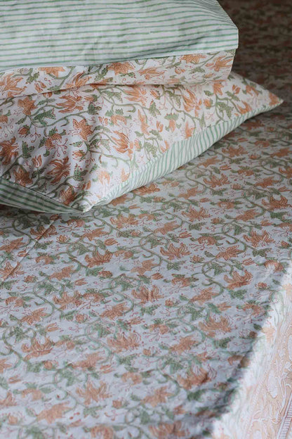 A close-up of a bed dressed in a floral print sheet and matching pillowcases. The pattern features orange flowers and green leaves on a light cream background, with a striped pillowcase in green and white at the top.