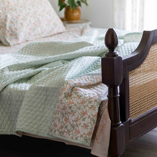 A quilted bedding set styled on a bed with a dark wood cane headboard. The set features a sage green and white striped quilt, a matching striped pillowcase, and a floral print quilt in soft pink, peach, and green tones.