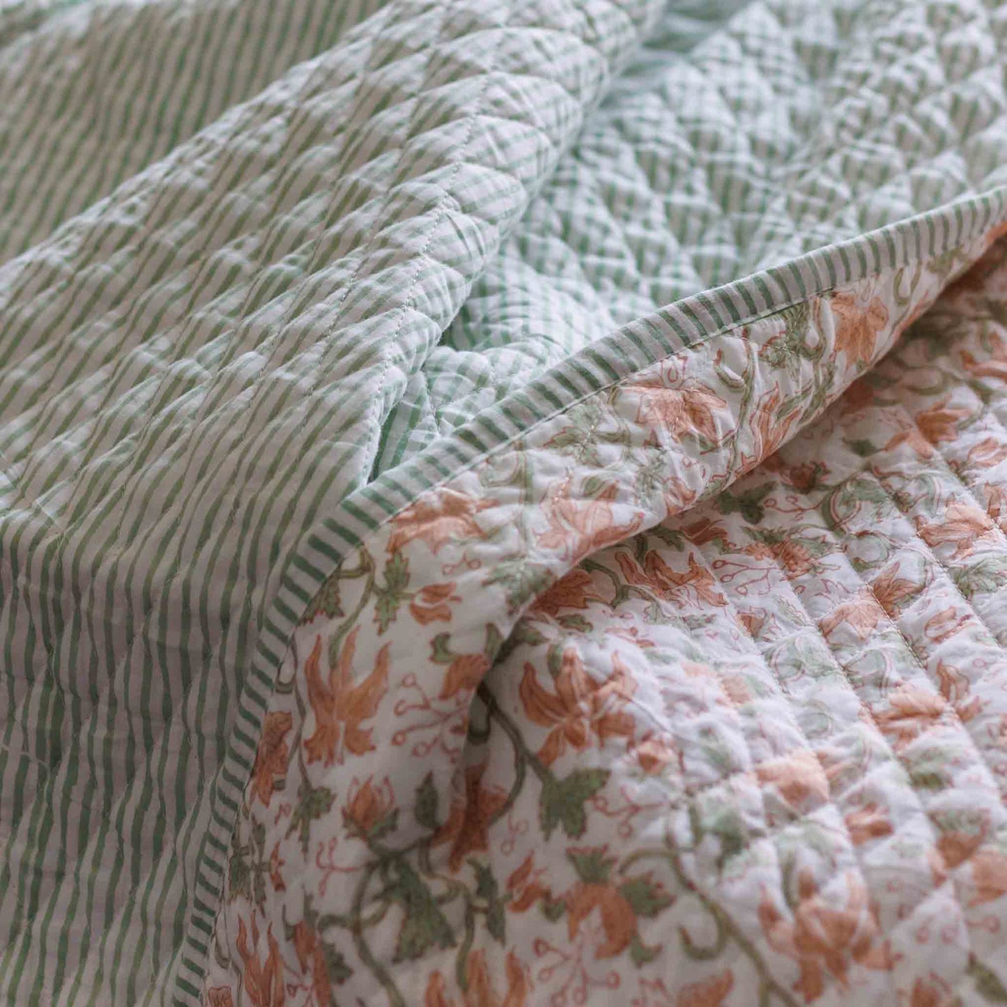 A close-up of a quilted blanket with a striped green and white side and a floral side in peach, green, and white, folded over to show its texture and pattern.
