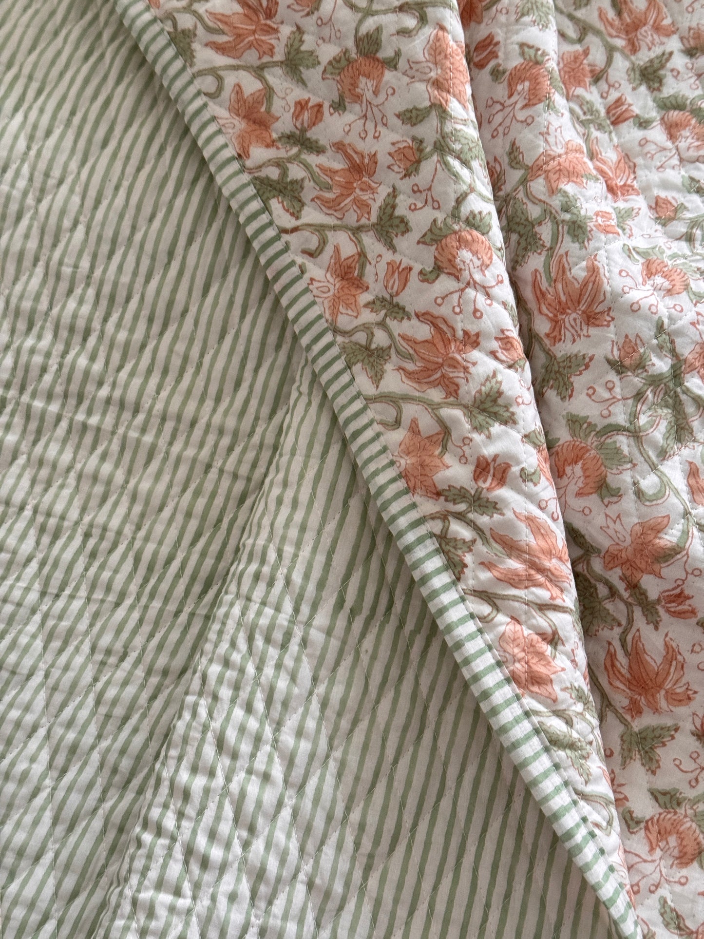Close-up of a quilted textile with two visible sides: one side shows fine vertical stripes in soft green and off-white, and the folded-over side shows a floral print in peach/coral and muted green on an off-white background.