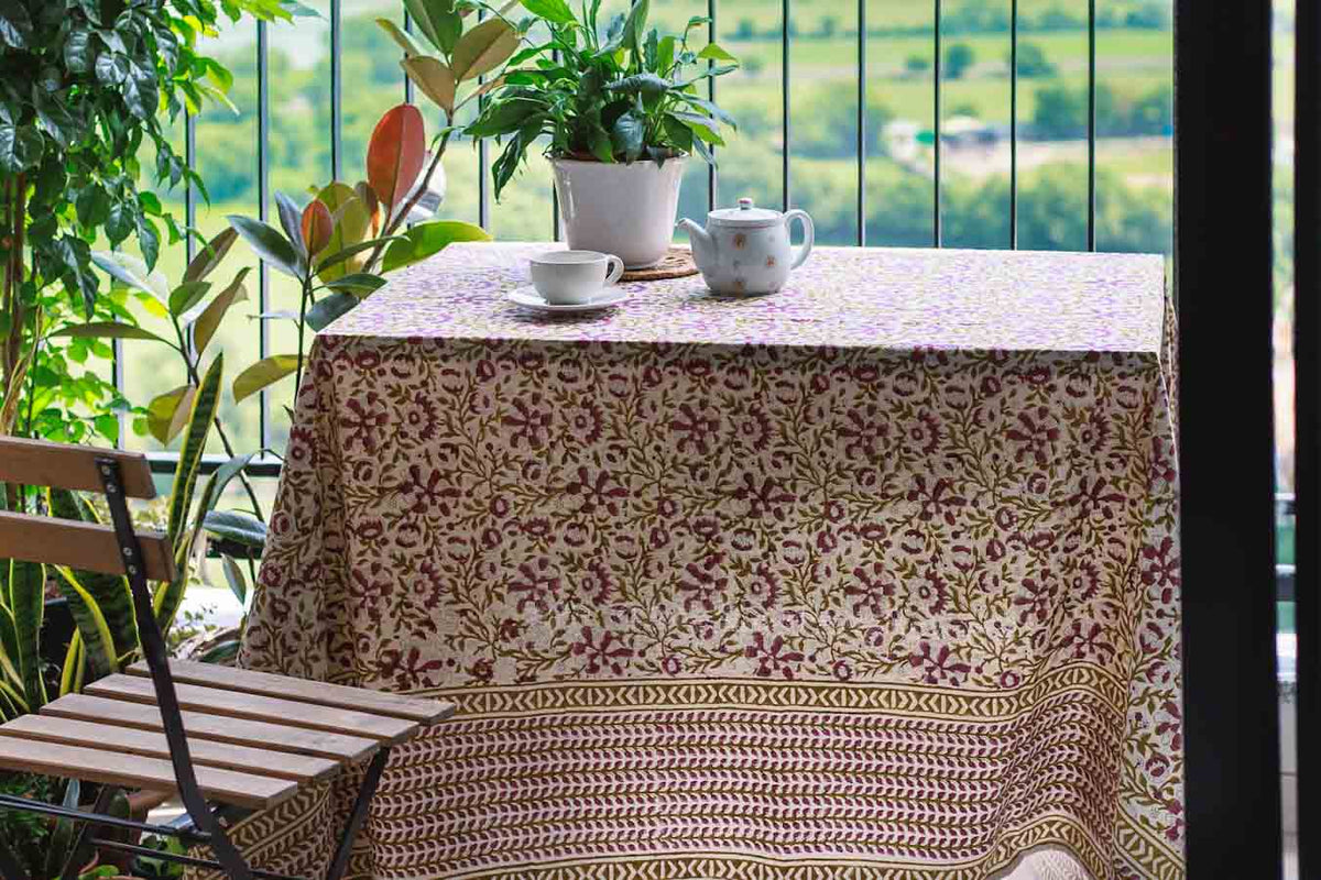 A rectangular table covered with tablecloth featuring a burgundy floral pattern and a geometric border, styled with a teapot, teacup, and potted plant on a balcony 
