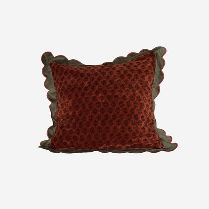 A square decorative throw pillow in rust red velvet with a subtle tonal floral pattern, featuring scalloped edges in a dark olive green velvet trim, shown against a plain light gray background.