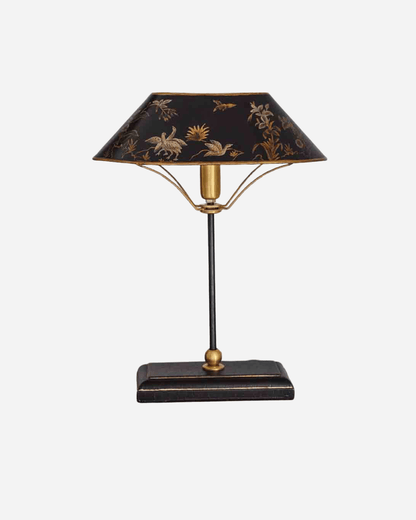 A vintage-style table lamp with a black shade decorated with gold floral motifs, supported by a gold-toned metal frame and a dark rectangular base.