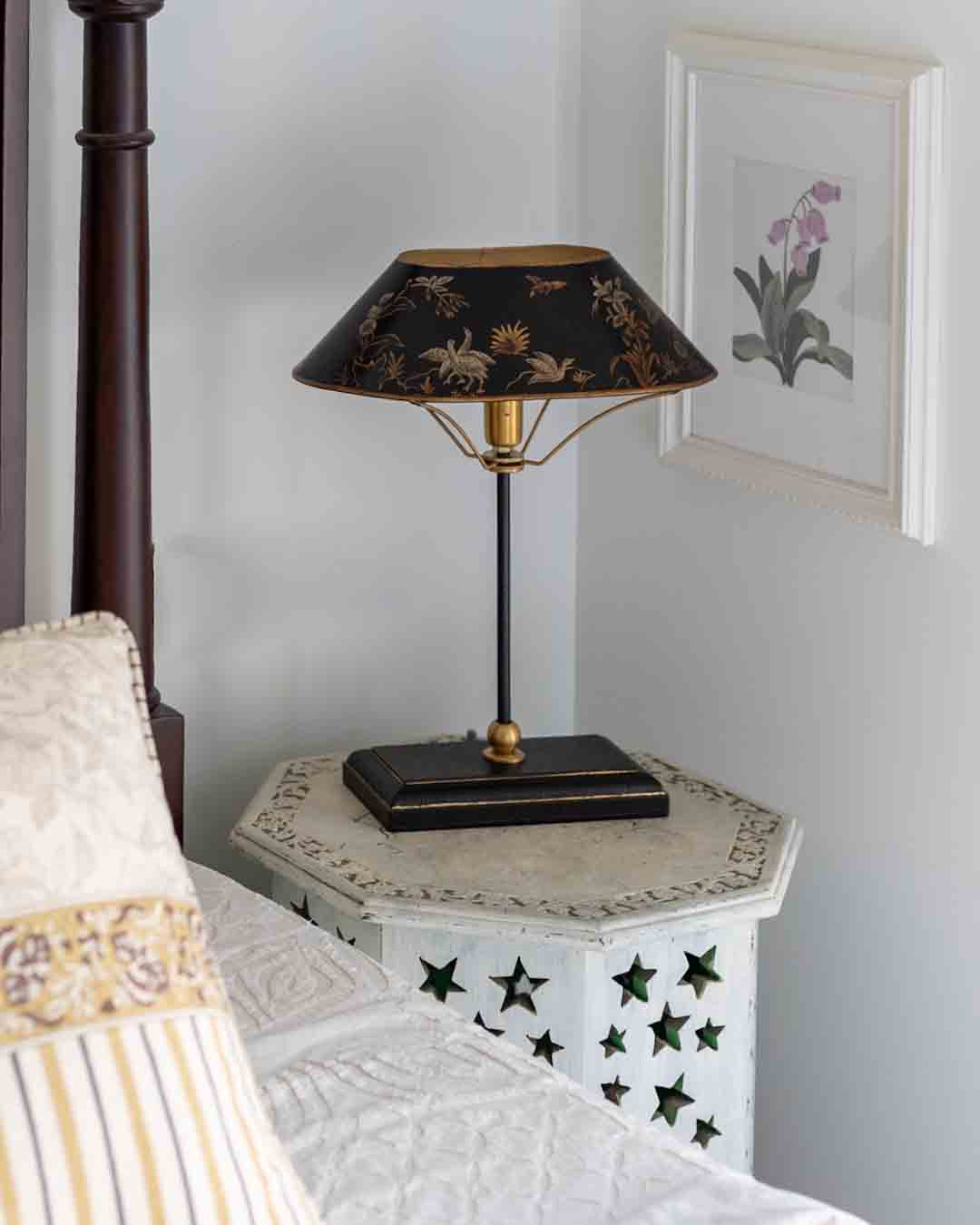 A vintage-style table lamp with a black shade decorated with gold floral and bird motifs, set on a white star-patterned nightstand in a bedroom.
