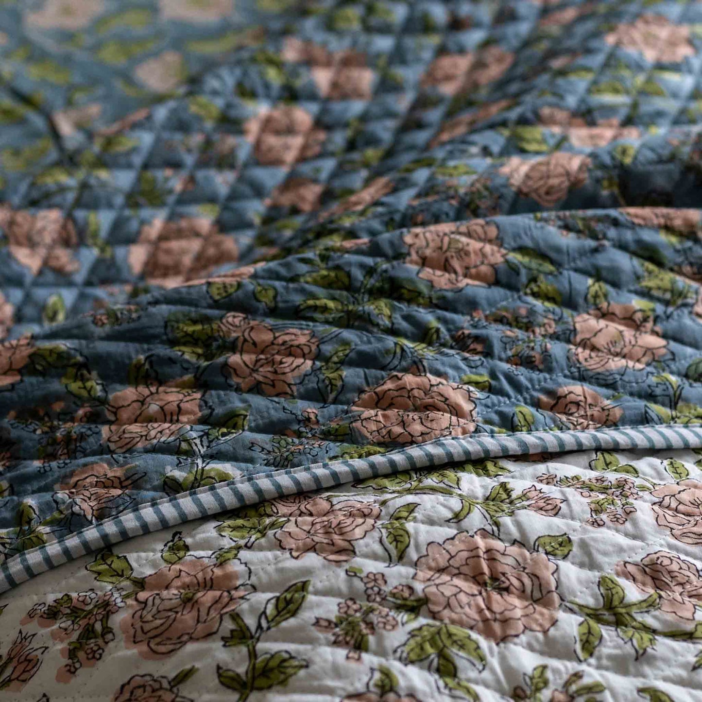 A close-up of a quilted textile with a floral print, showing a dusty blue top layer with pink roses and green leaves layered over a white bottom layer with a matching floral print and a blue-and-white gingham border.