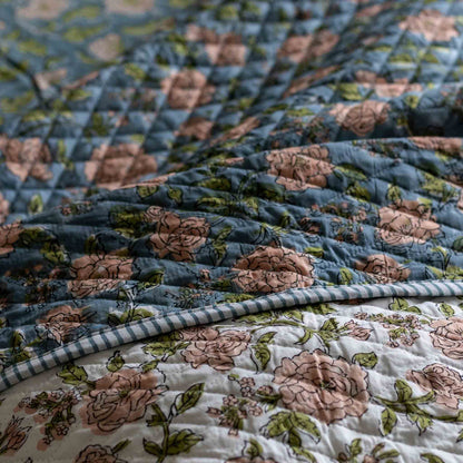 A close-up of a quilted textile with a floral print, showing a dusty blue top layer with pink roses and green leaves layered over a white bottom layer with a matching floral print and a blue-and-white gingham border.
