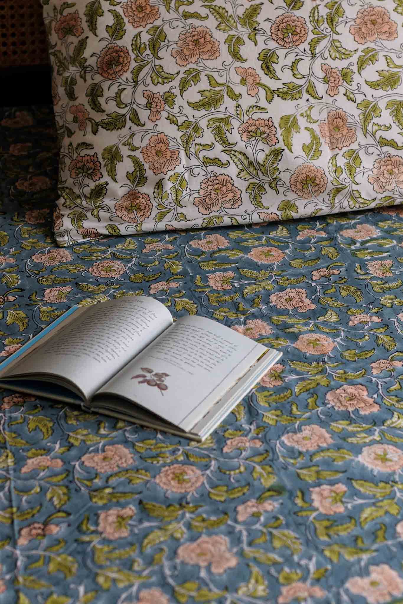 A close-up of a blue floral quilt or bedspread with a matching pillow, featuring an open book on the surface.