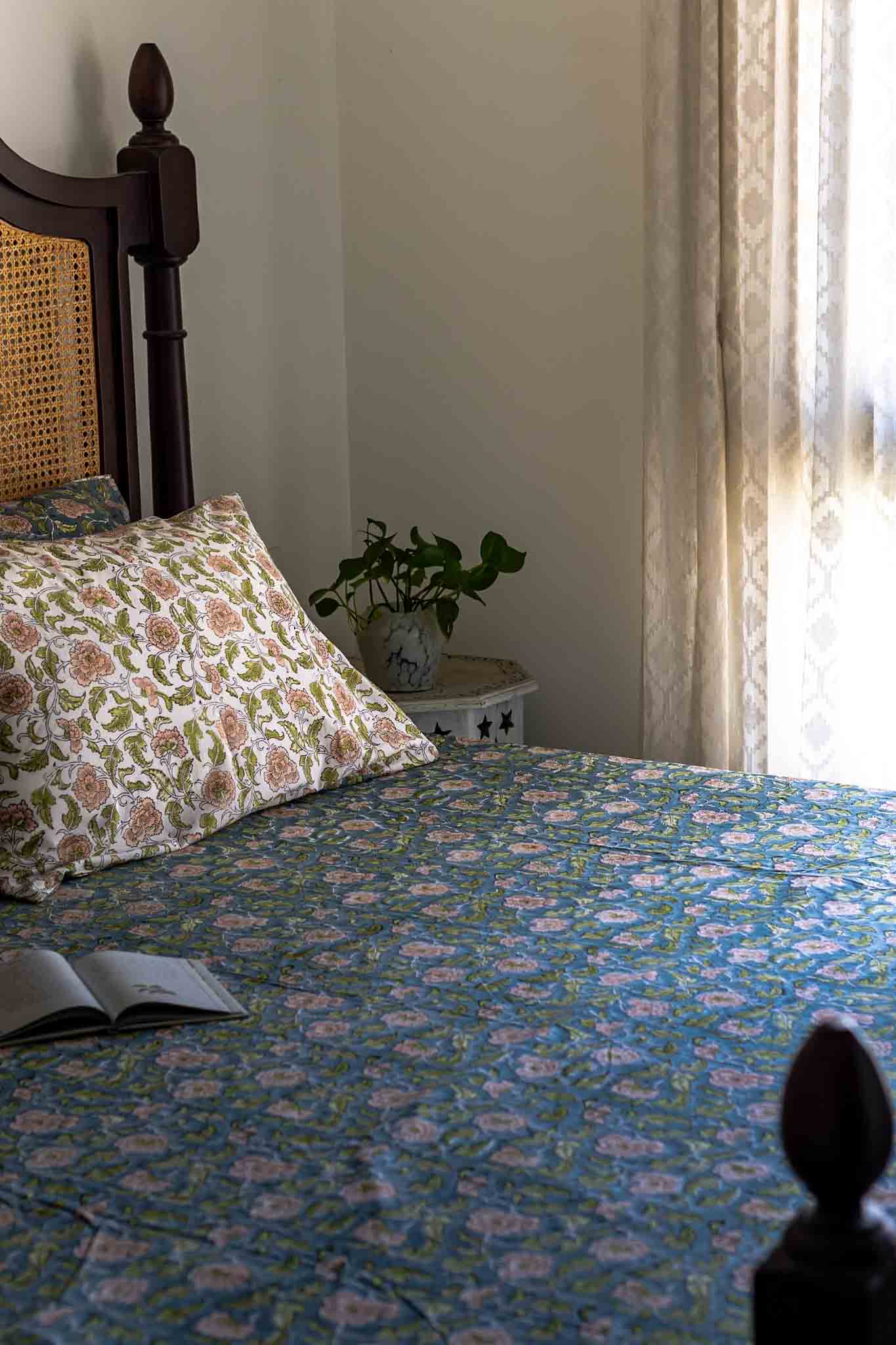 A bed dressed in a blue floral patterned bedspread and matching pillow shams, styled in a softly lit bedroom with a dark wooden headboard, curtain, small side table, and an open book on the bed.