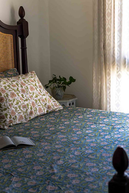 A bed dressed in a blue floral patterned bedspread and matching pillow shams, styled in a softly lit bedroom with a dark wooden headboard, curtain, small side table, and an open book on the bed.