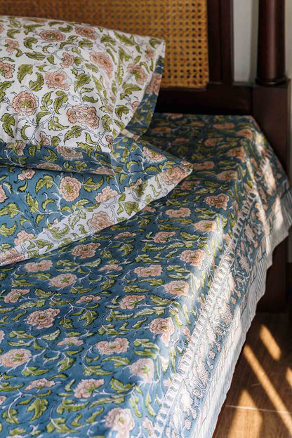 A close-up of a bed dressed in floral bedding, featuring a blue duvet cover and matching pillowcases with pink and green botanical patterns, styled in a warm, sunlit bedroom setting.