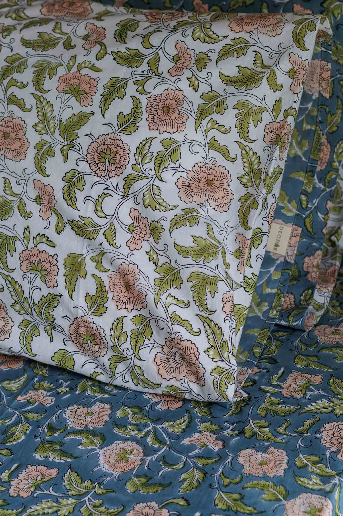 A close-up of a floral-patterned bedding set, featuring a white pillowcase with pink flowers and green leaves layered over a blue duvet cover with the same floral print.