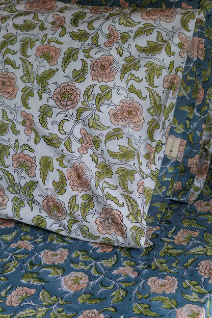 A close-up of a floral-patterned bedding set, featuring a white pillowcase with pink flowers and green leaves layered over a blue duvet cover with the same floral print.