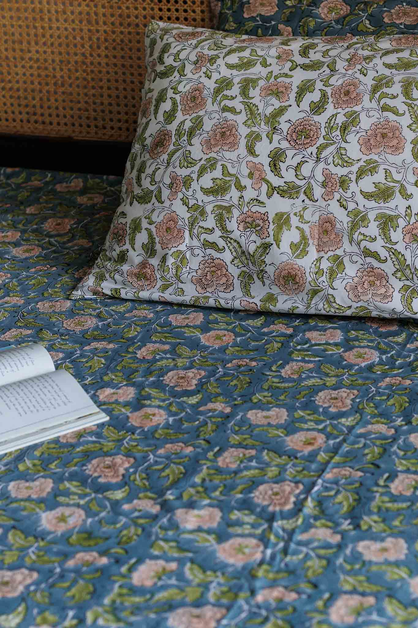 A close-up of a bed styled with a blue floral quilt and matching pillowcase, featuring pink and green botanical patterns. An open book lies on the bed next to a woven cane headboard.
